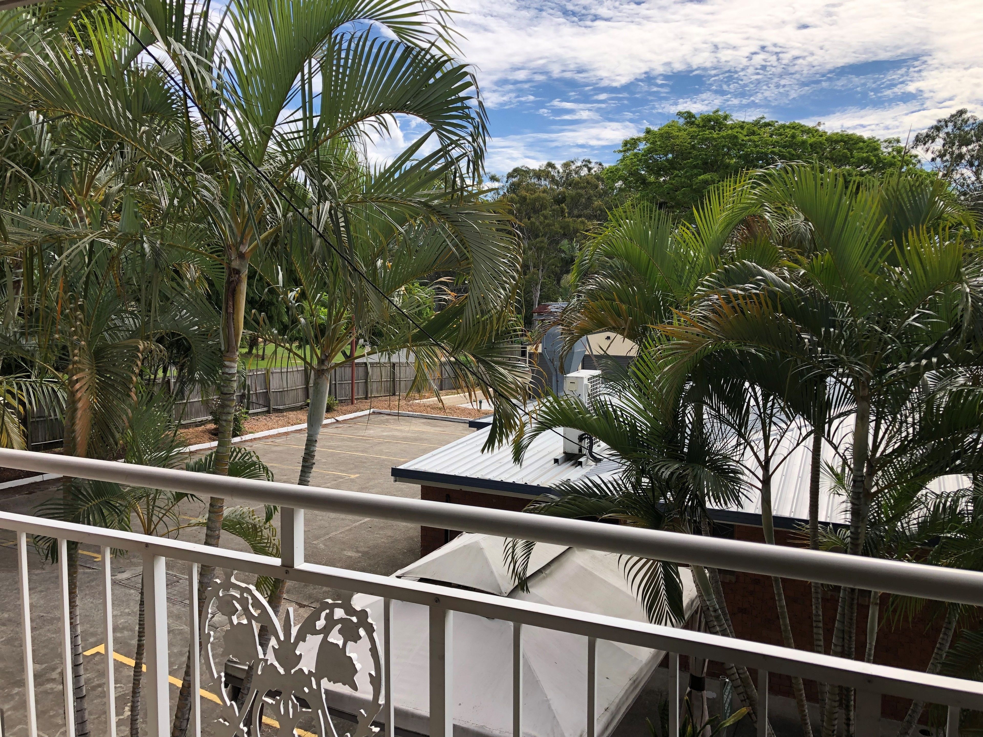 A balcony with a view of a parking area, palm trees, and a building with a metal roof.
