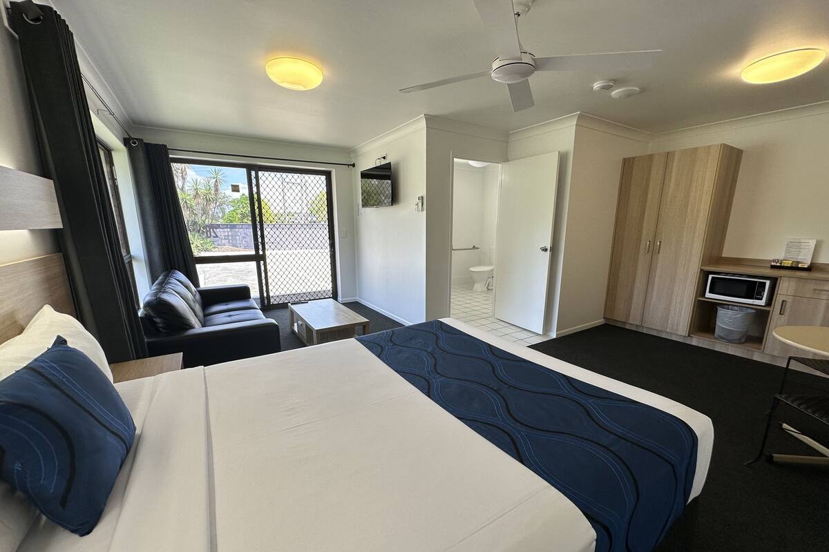 A hotel room with a bed, a small table, a sofa, a kitchenette, and a bathroom visible through an open door.