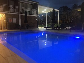 A swimming pool with a clear blue water surface, surrounded by lounge chairs and a covered area with a glass wall.
