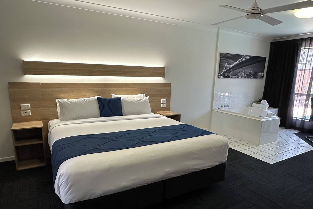 A modern hotel room with a large bed, bedside tables, a ceiling fan, a bathroom with a bathtub, and a cityscape view.
