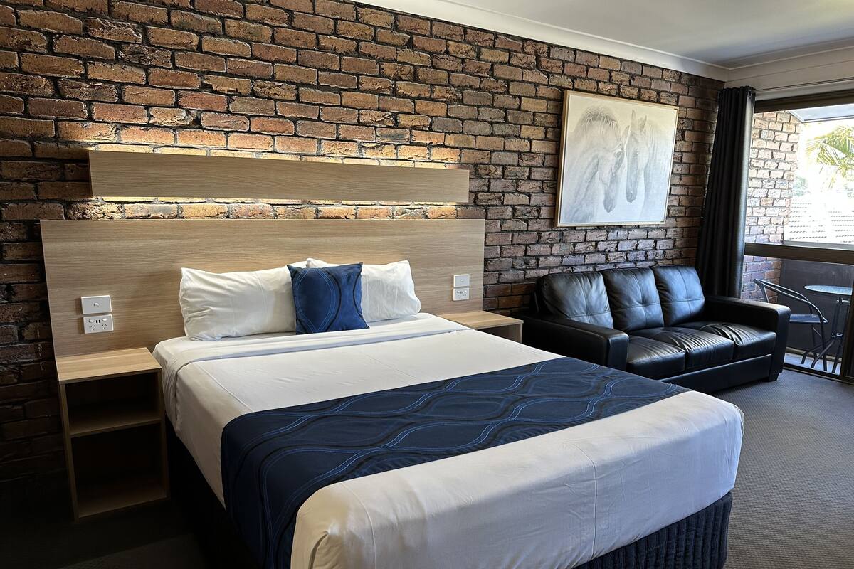 A hotel room with a brick wall, a large bed with a blue quilt, a leather sofa, and a painting above the sofa.