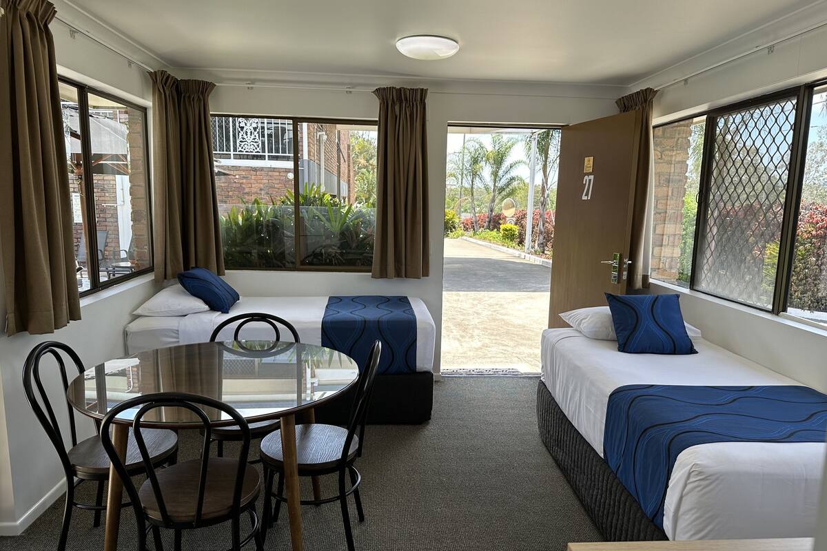 A hotel room with two beds, a dining table with chairs, and large windows with curtains.