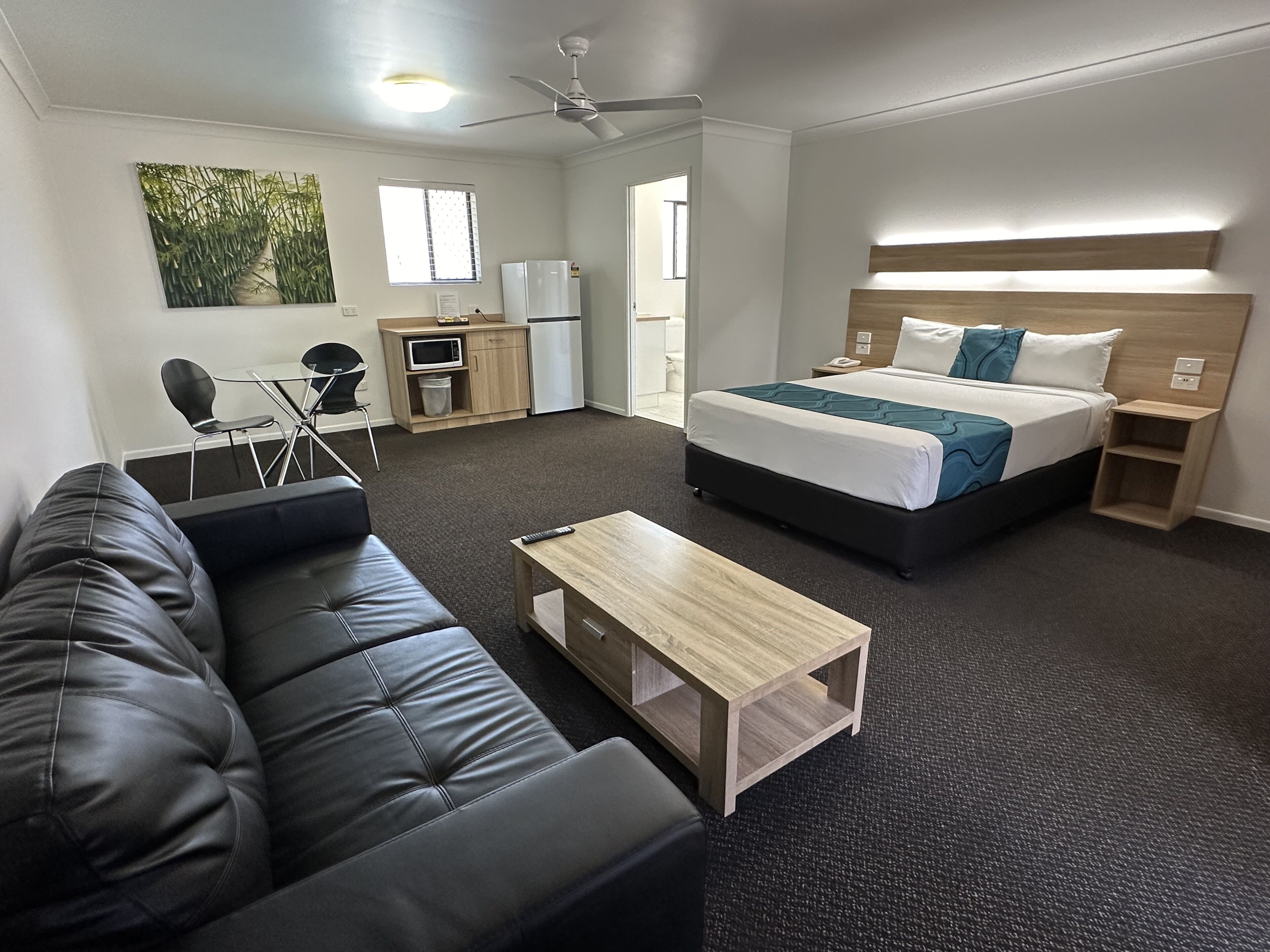Una habitación de hotel moderna con sofá de cuero negro, una mesa de centro de madera, una cama con cabecera de madera, un escritorio pequeño con una silla, un microondas y un refrigerador.