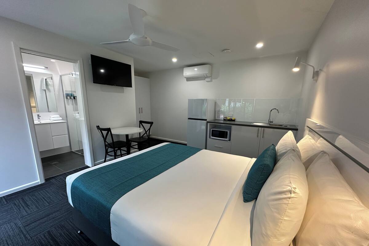 A hotel room with a bed, a desk, a chair, a TV, a ceiling fan, and a kitchenette.