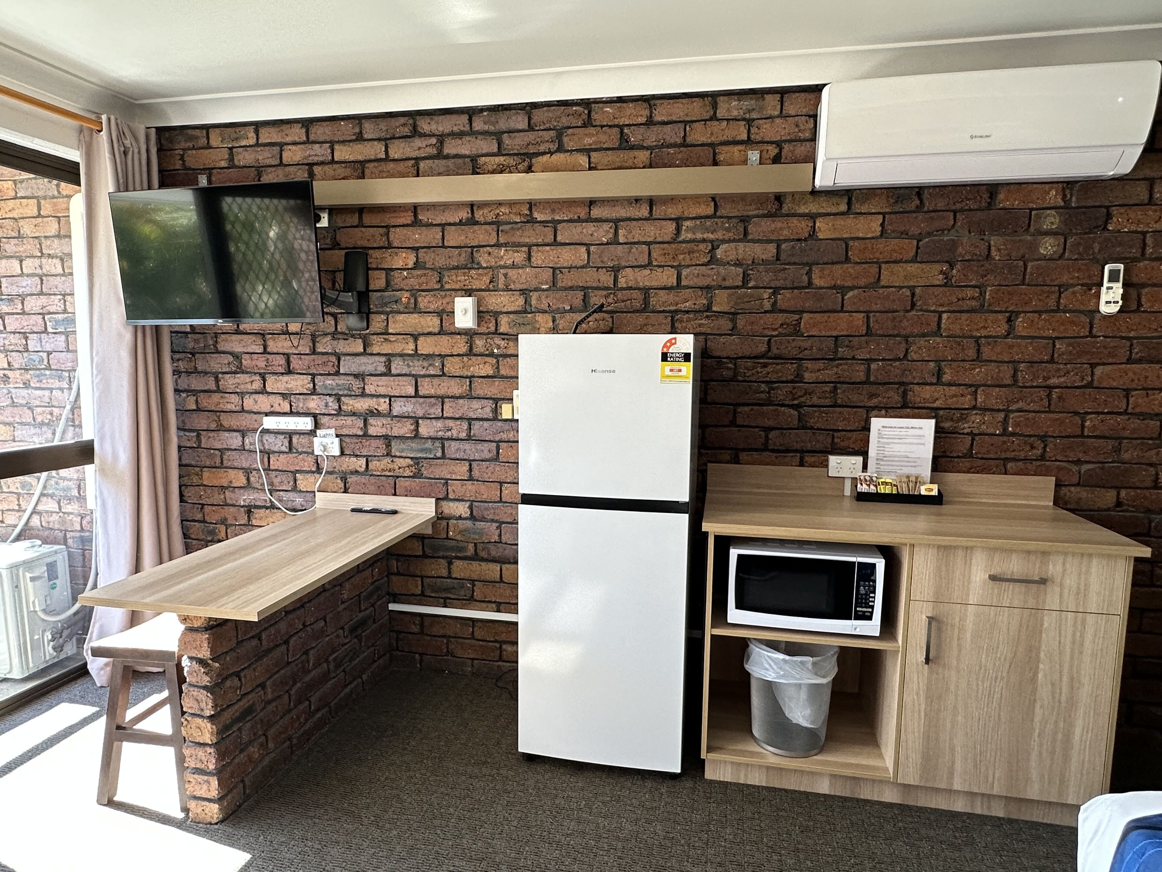 Cocina compacta con pared de ladrillo, refrigerador, microondas, mesa de madera y televisor montado en la pared.
