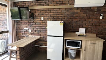 Cocina compacta con pared de ladrillo, refrigerador, microondas, mesa de madera y televisor montado en la pared.