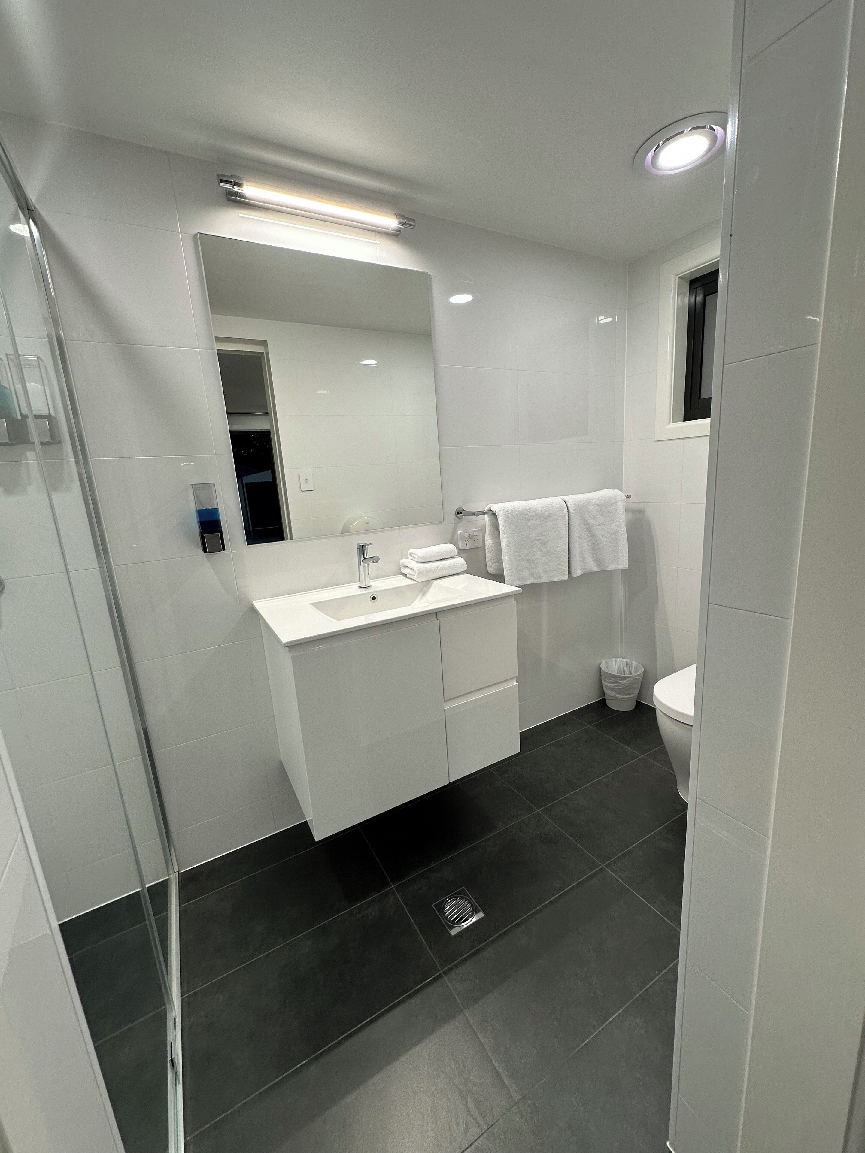 A modern bathroom with a white vanity, a large mirror, a toilet, and a shower area.