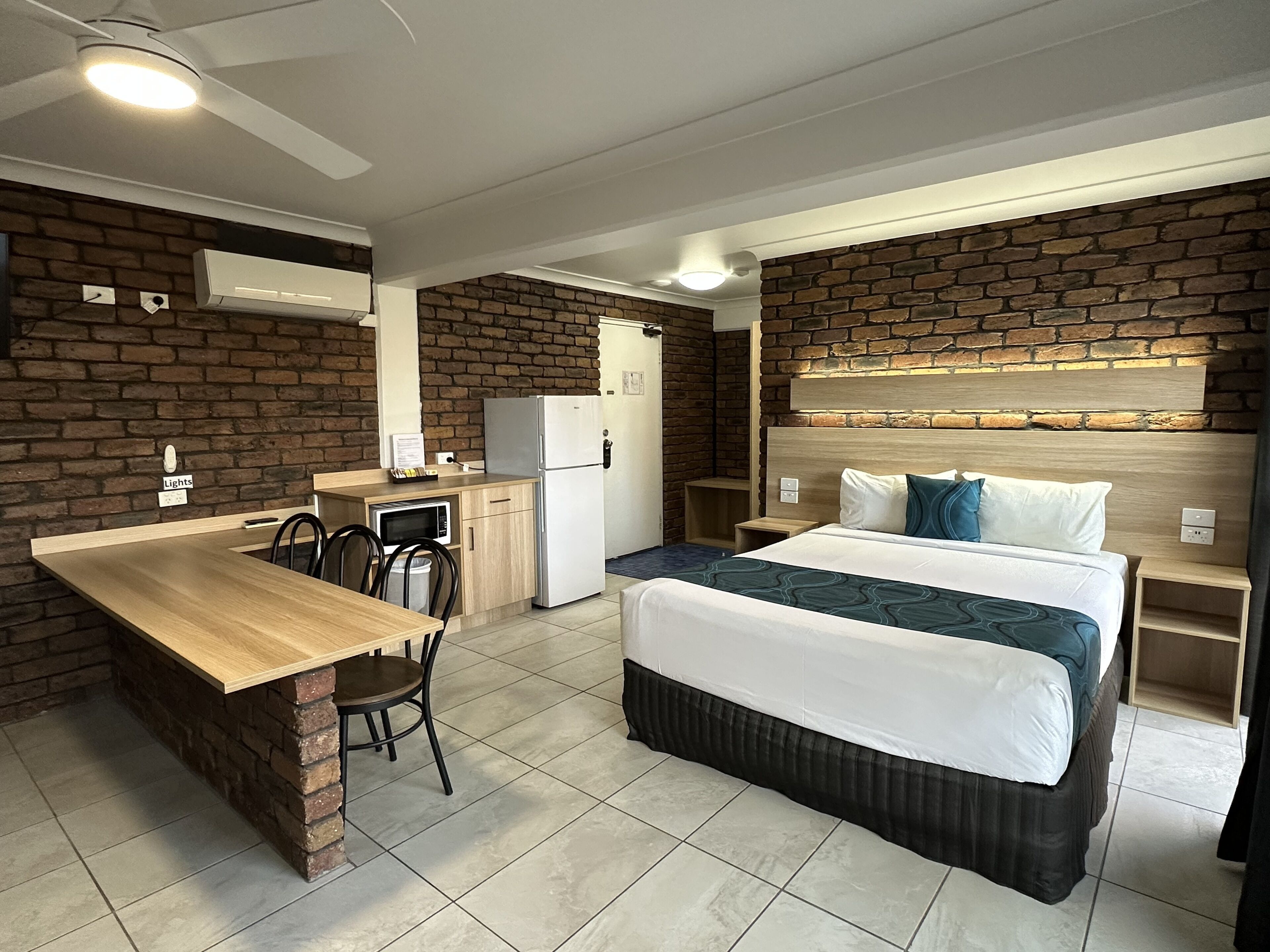 A compact hotel room with a kitchenette, a bed with a headboard, a dining area with a table and chairs, and a brick accent wall.