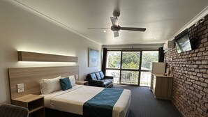 A hotel room with a bed, a desk, a chair, a sofa, a ceiling fan, a TV, and a large window with a view of greenery.