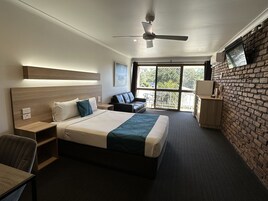 Habitación de hotel con cama, escritorio, silla, sofá, ventilador de techo, televisor y una ventana grande con vistas a zonas verdes.