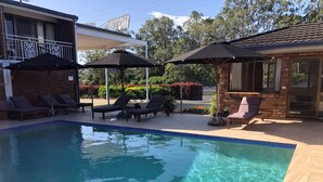 A swimming pool with clear water, surrounded by lounge chairs and umbrellas, and a building with a balcony in the background.