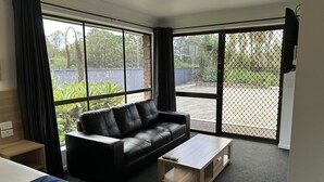 A modern hotel room with a black leather sofa, a wooden coffee table, and a large window offering a view of the outdoors.
