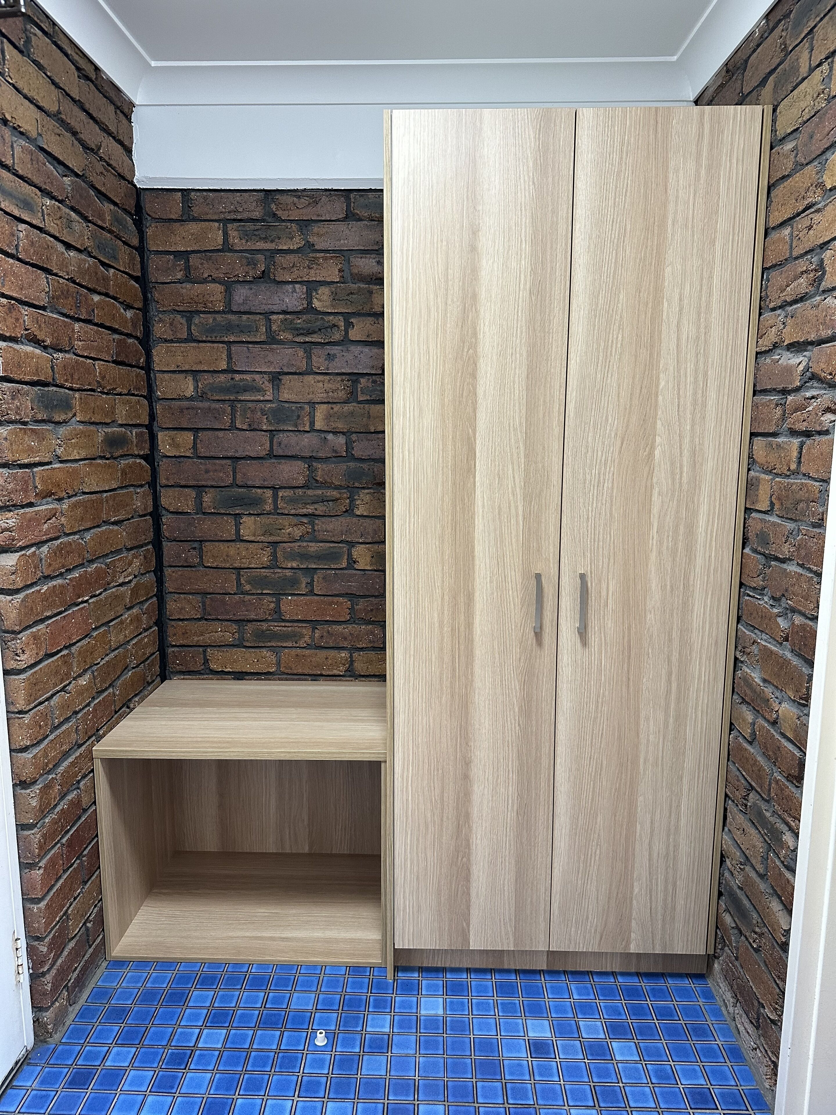 A room with a brick wall, a wooden bench, and a wardrobe with two doors.