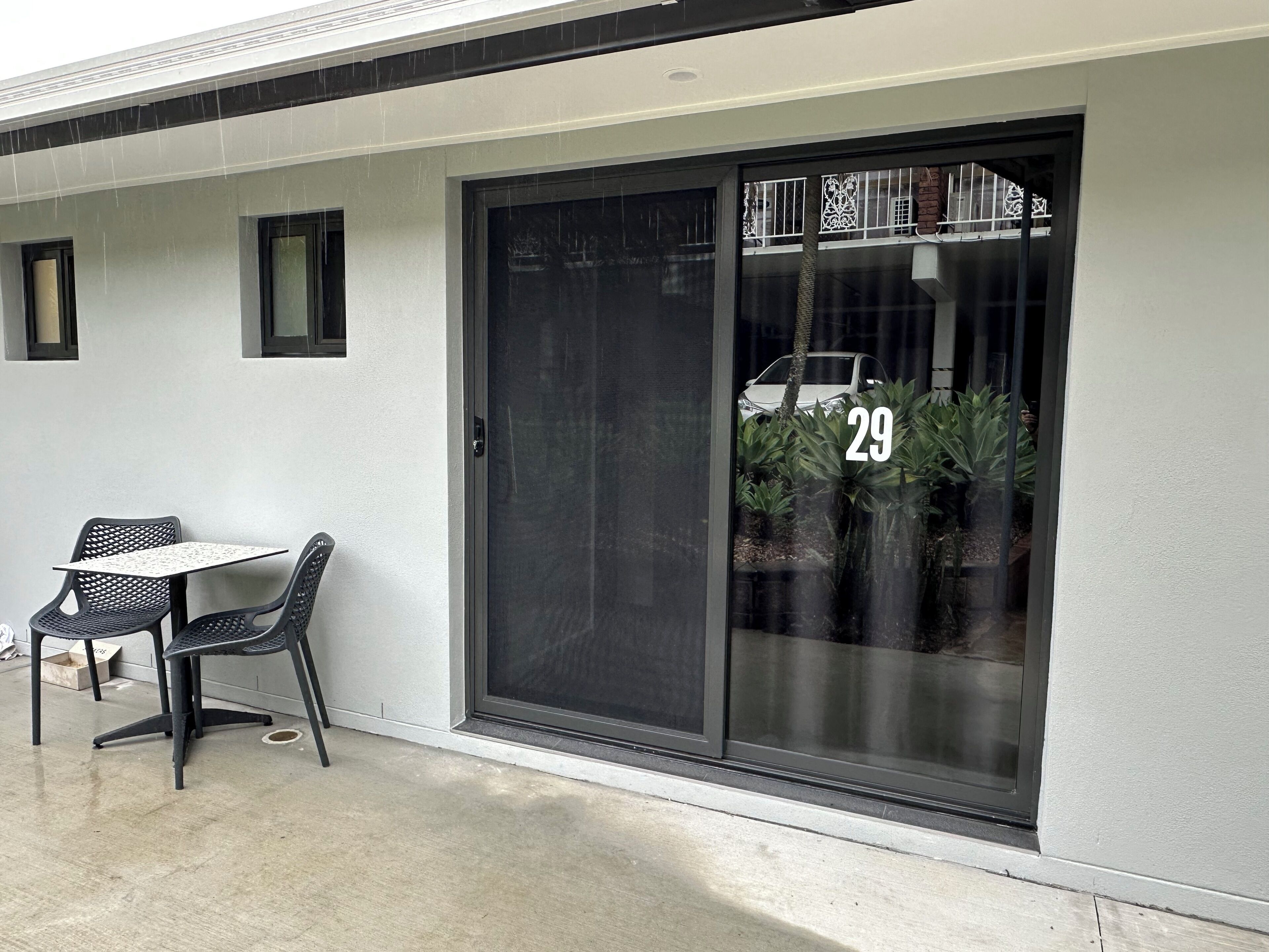 A modern building with a large sliding glass door marked with the number 29, a small outdoor table with chairs, and a concrete floor.