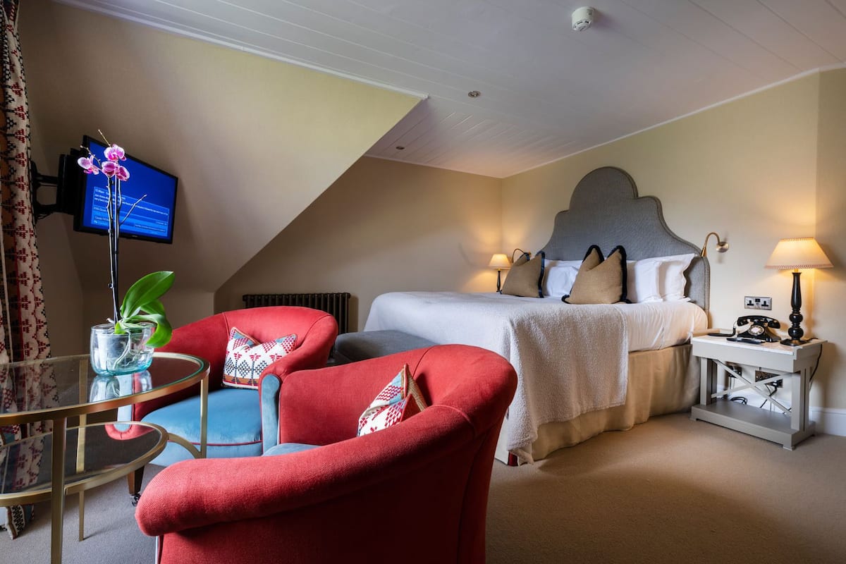 A hotel room with a bed, two red armchairs, a glass table with a plant, a TV, and a bedside table with a lamp.