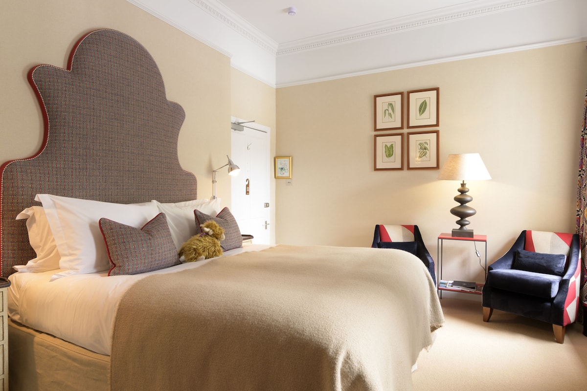 A hotel room with a large bed, a chair, a lamp, and framed botanical prints on the wall.