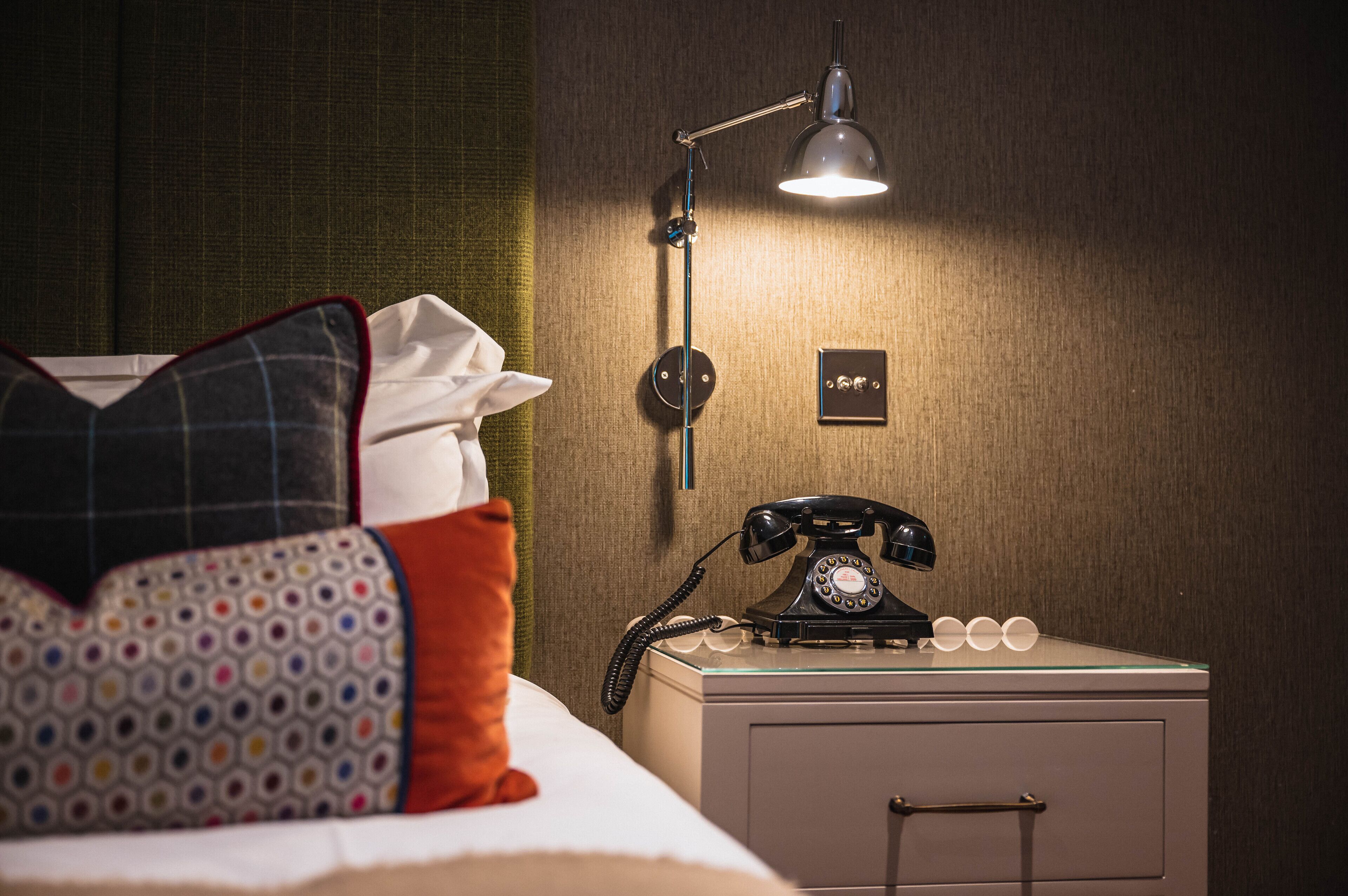A hotel room with a bed, pillows, a bedside table with a rotary phone, a lamp, and a wall-mounted light switch.