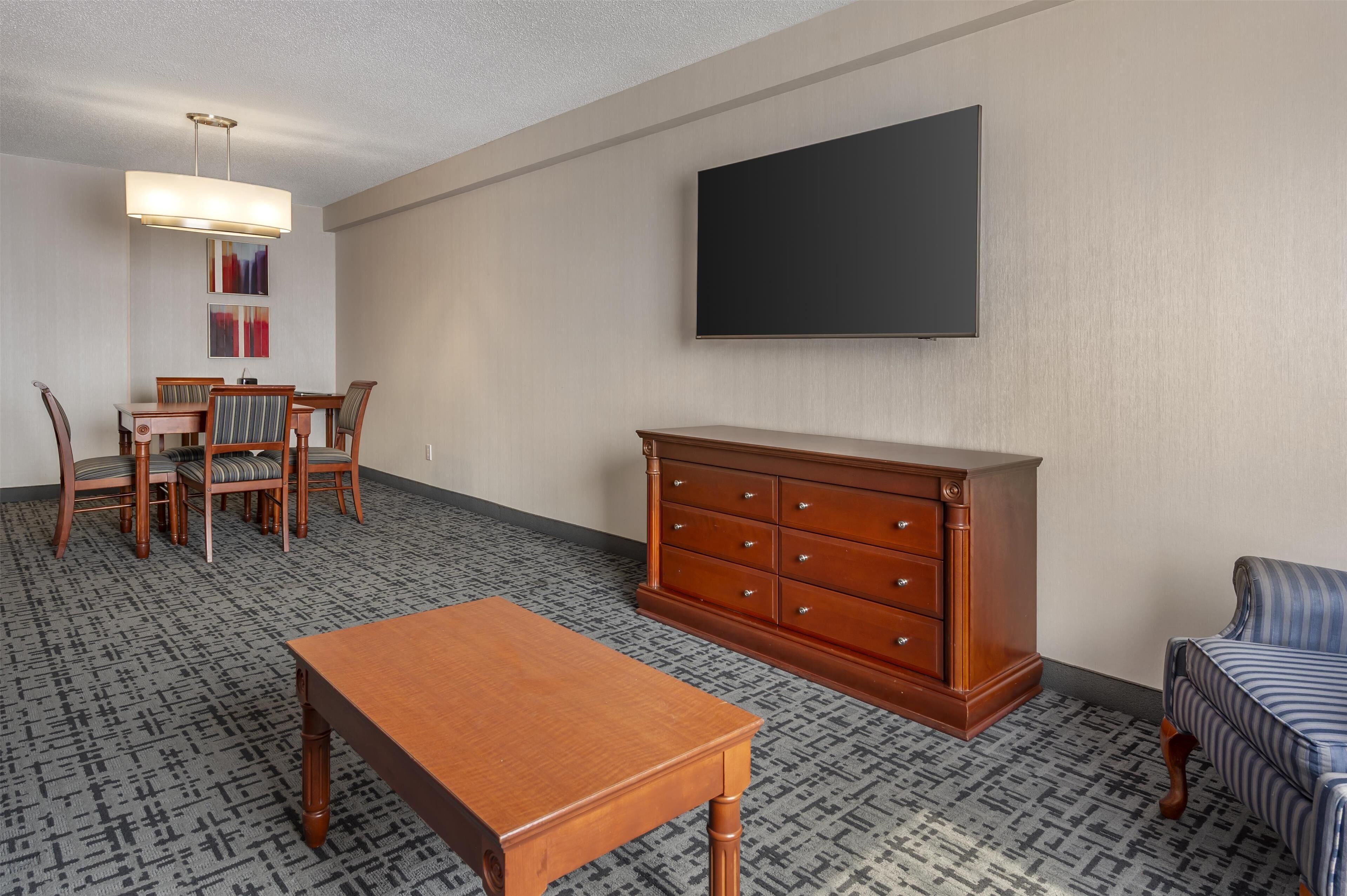 Une chambre d’hôtel équipée d’une table à manger, de chaises, d’un téléviseur fixé au mur, d’une commode en bois et d’un tapis à motifs.