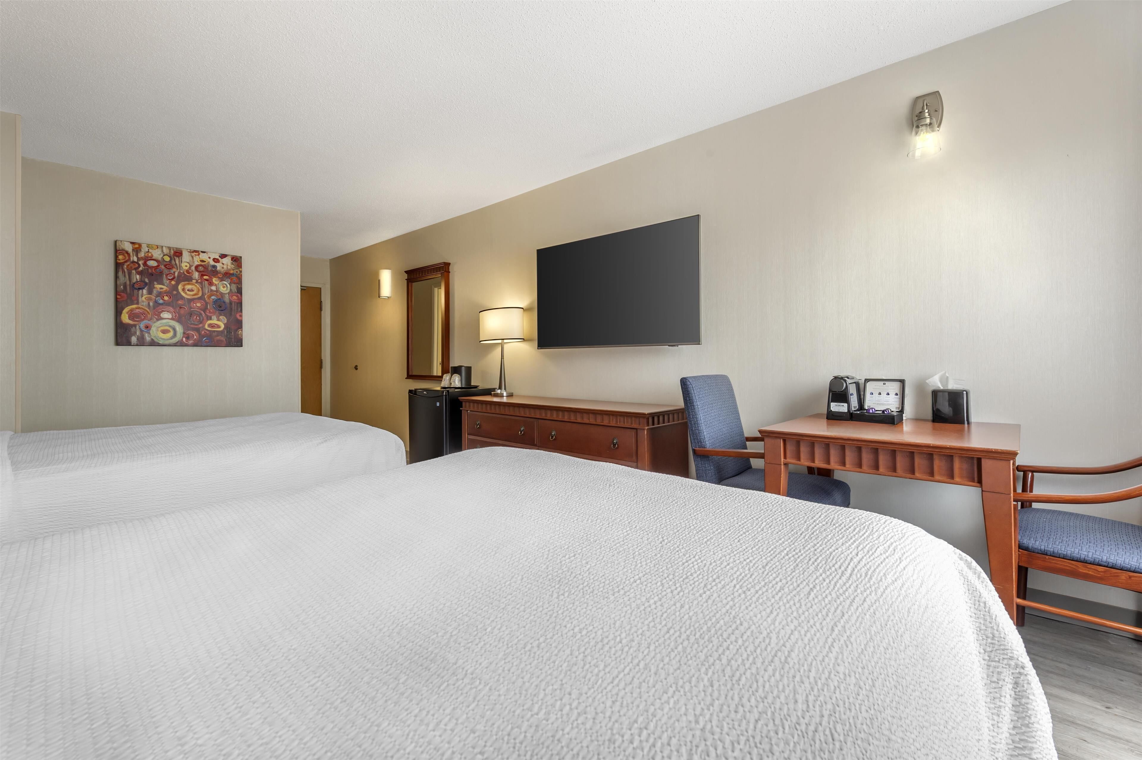 Habitación de hotel con dos camas, un escritorio con silla, televisor de pantalla plana, lámpara de escritorio y un mueble pequeño.