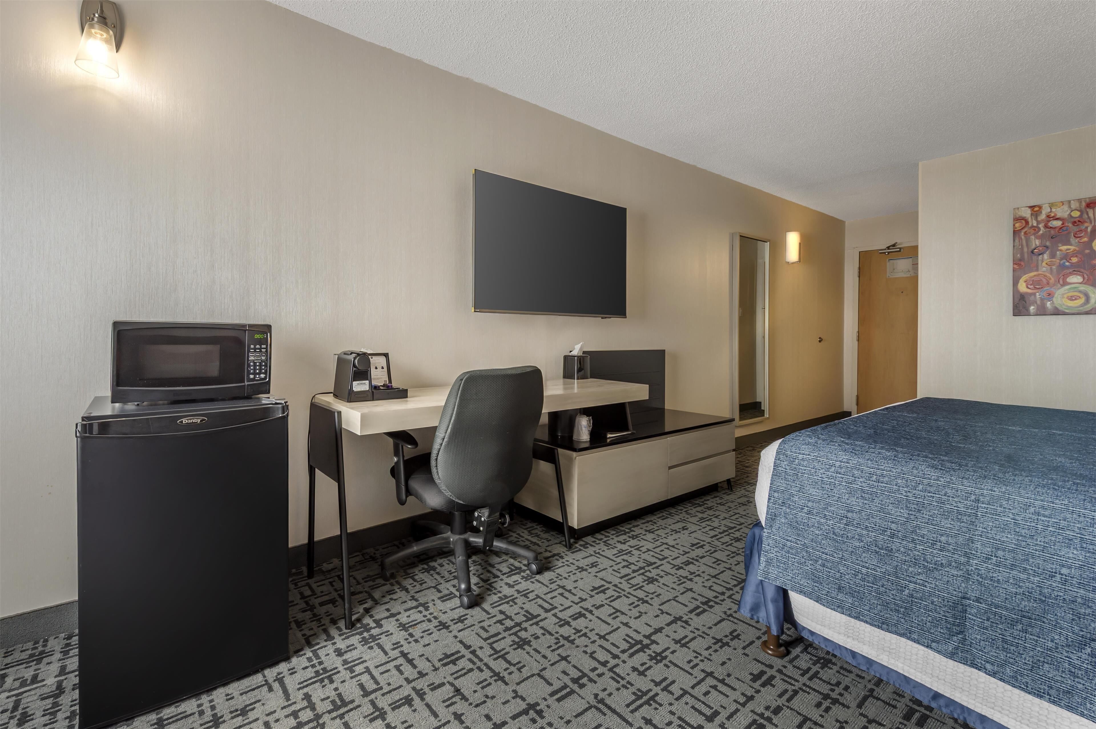A hotel room with a bed, a desk with a computer, a microwave, and a refrigerator.