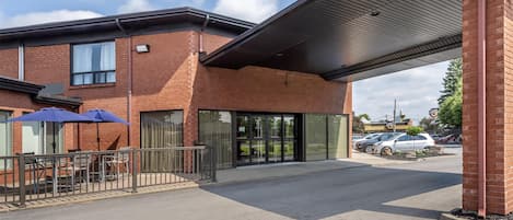 Un bâtiment moderne doté d’une entrée abritée, de grandes fenêtres et d’un espace de stationnement où sont garées des voitures.