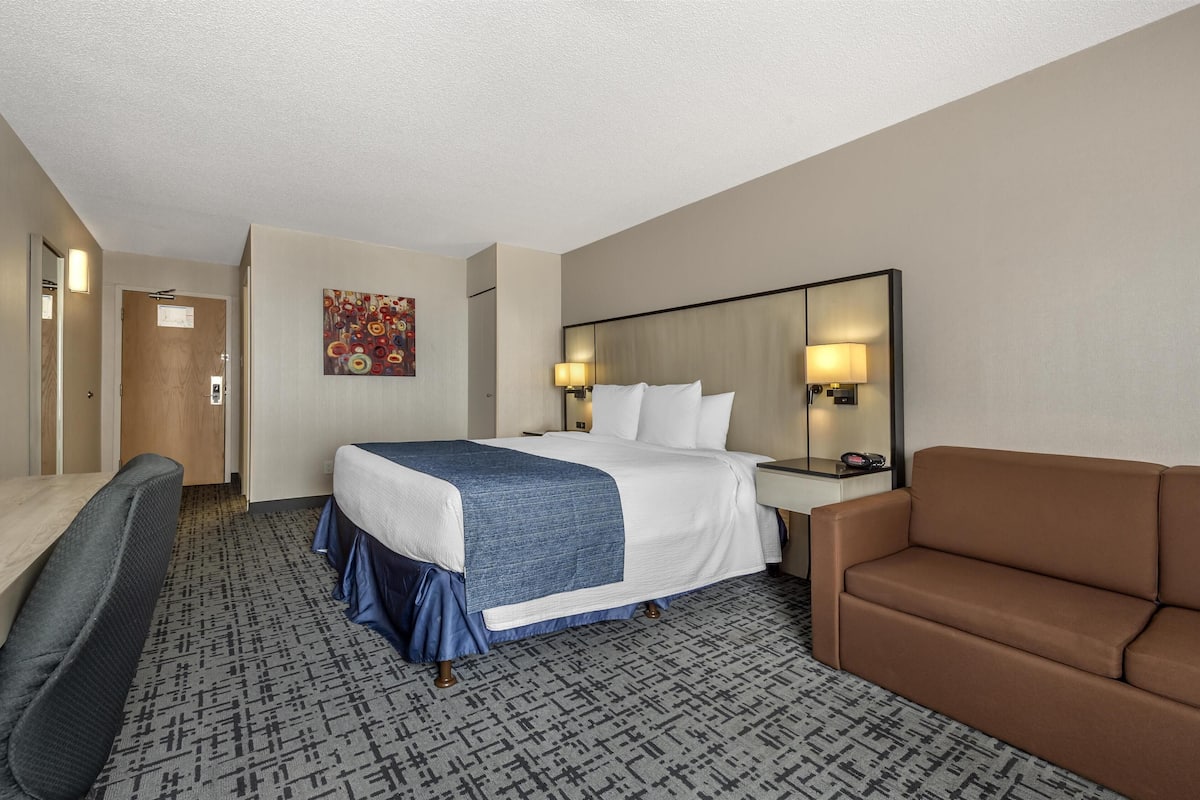 A hotel room with a bed, a desk, a chair, a sofa, and a painting on the wall.