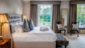 A neatly made bed with a tufted headboard, a bedside table with a lamp, and a window with curtains. - Sudbury House Hotel (Faringdon)