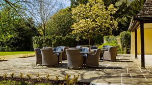An outdoor patio with wicker furniture and a stone floor, surrounded by greenery and trees. - Sudbury House Hotel (Faringdon)