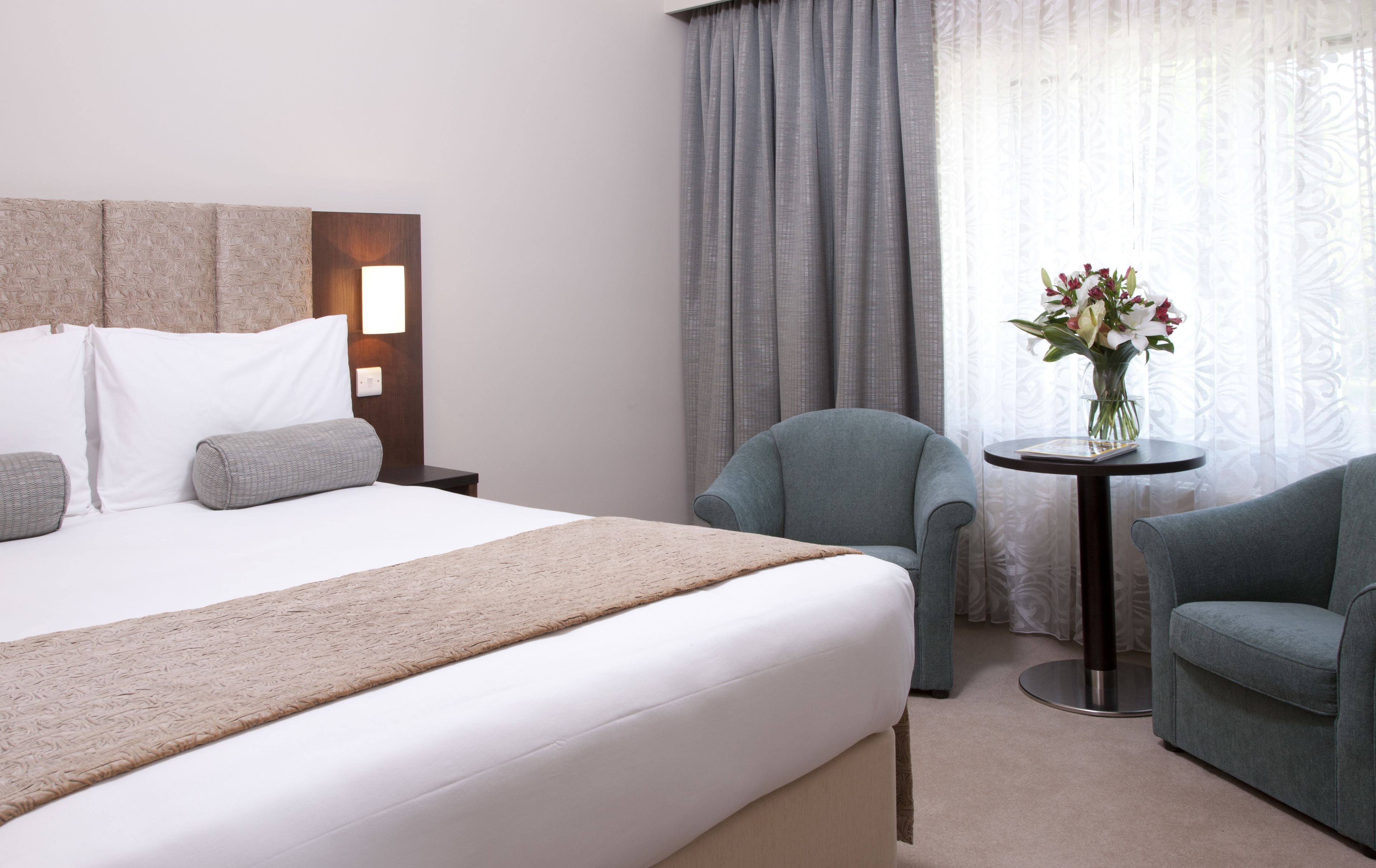 Habitació d'hotel amb un llit gran, dos seients, una taula petita amb un ram de flors i una finestra amb cortines.