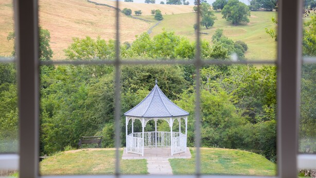 A gazebo with a white railing and a bench, surrounded by lush greenery and hills in the background. - The Izaak Walton Hotel (Ashbourne)