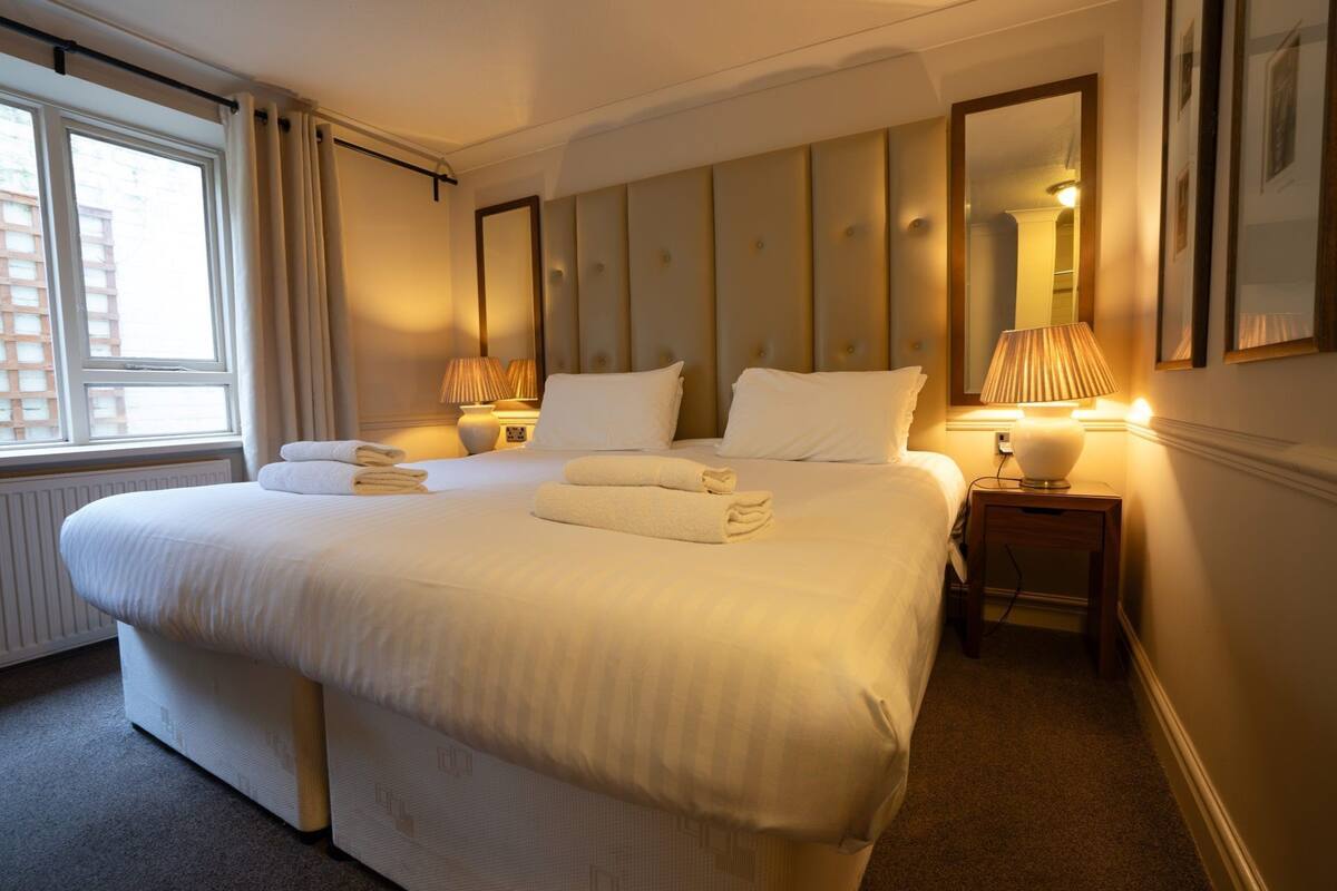 A large bed with white linens and towels, a headboard with vertical panels, two bedside lamps, and a large mirror on the wall.