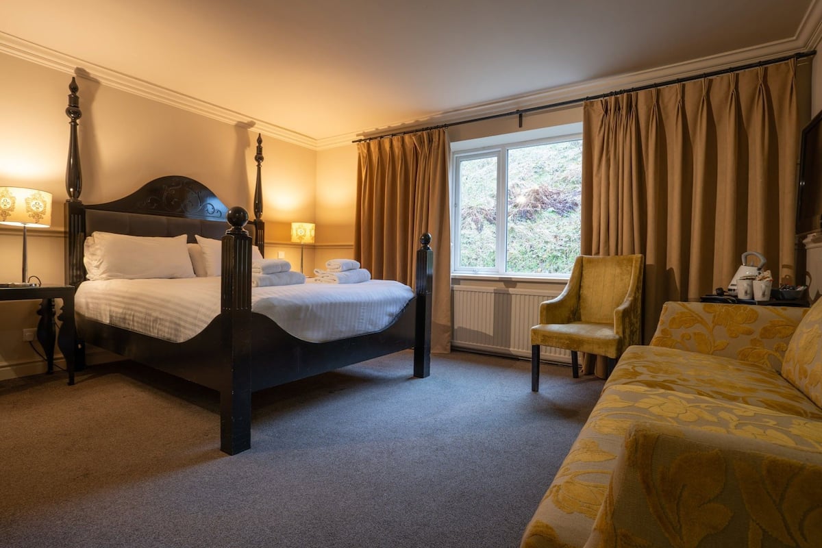 A four-poster bed with a dark finish, a sitting chair, and a sofa with a floral pattern.