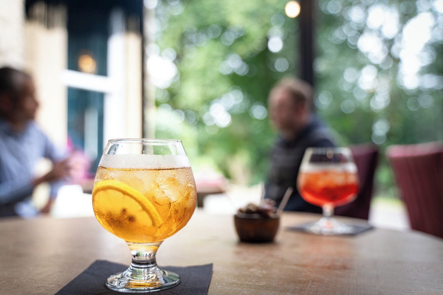 A glass of beer with a lemon slice on a table in a restaurant with a view of greenery outside.