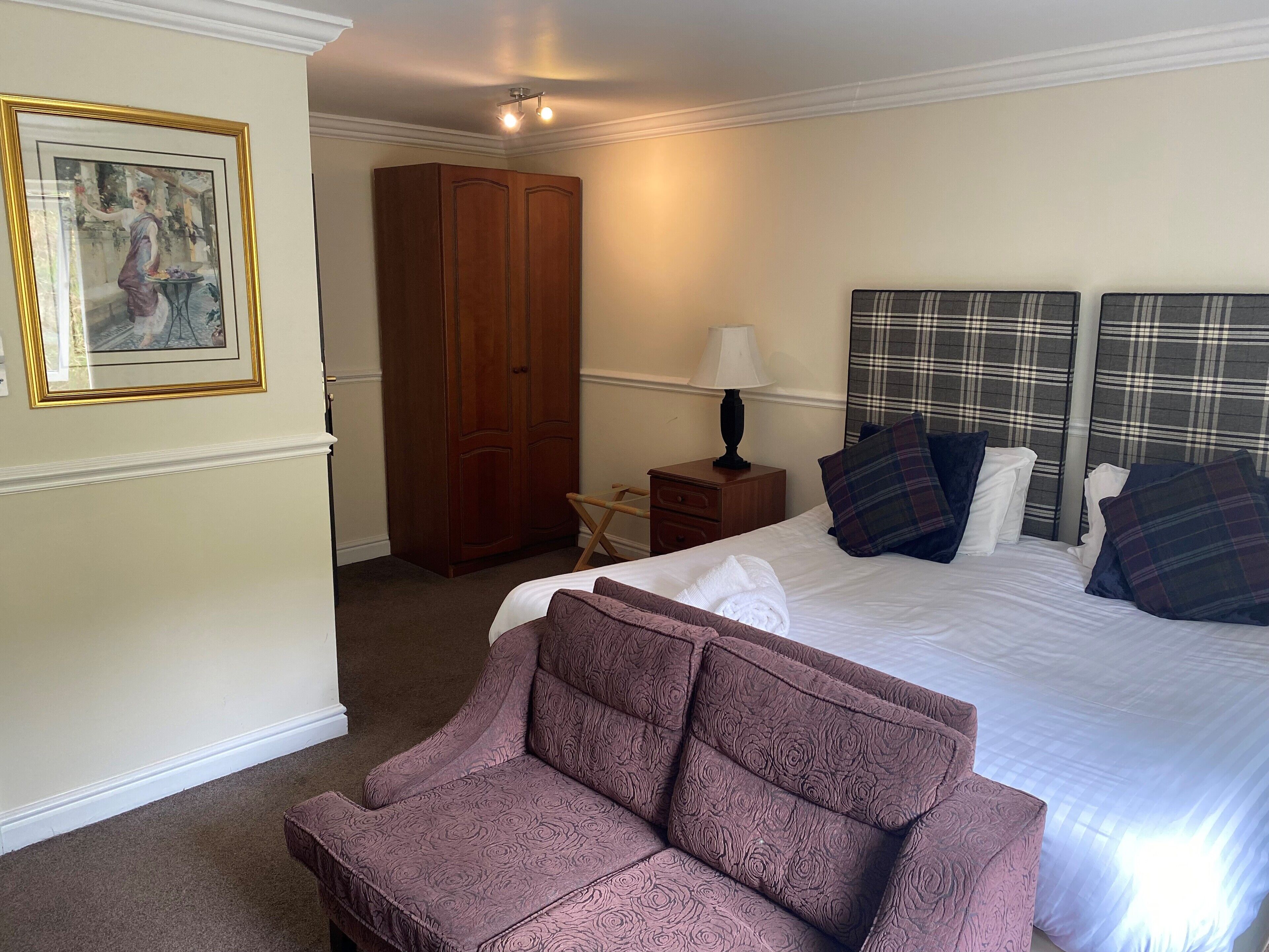 Ein Hotelzimmer mit einem großen Bett, einem hölzernen Kleiderschrank, einem kleinen Tisch mit Lampe und einem gerahmten Bild an der Wand.