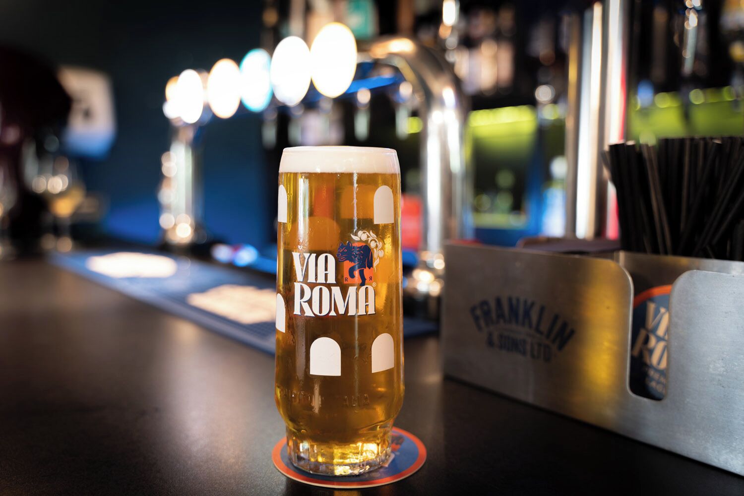 A glass of beer with "VIA ROMA" on it, placed on a coaster.