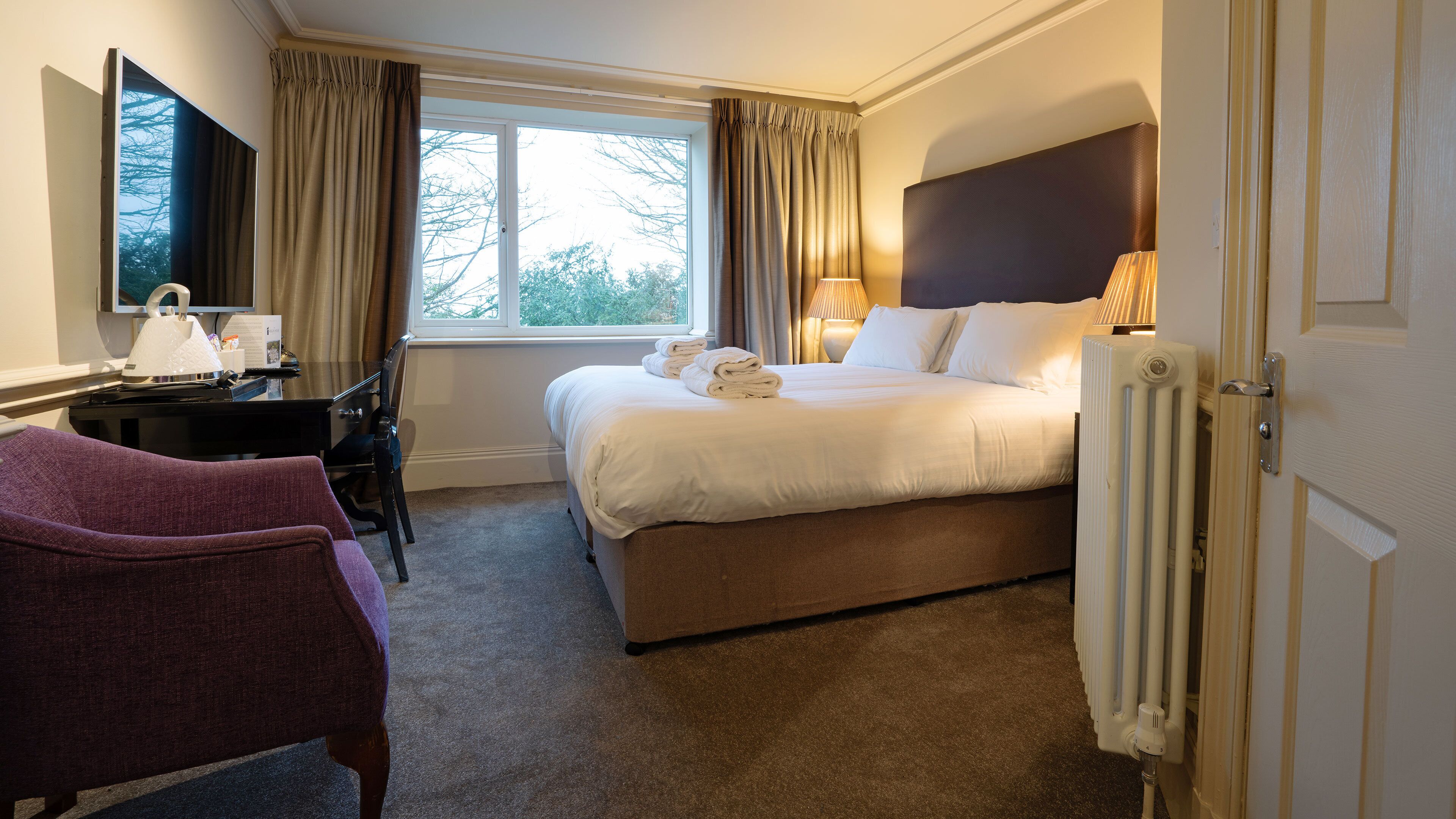 A hotel room with a bed, a desk, a chair, a window with curtains, and a radiator.