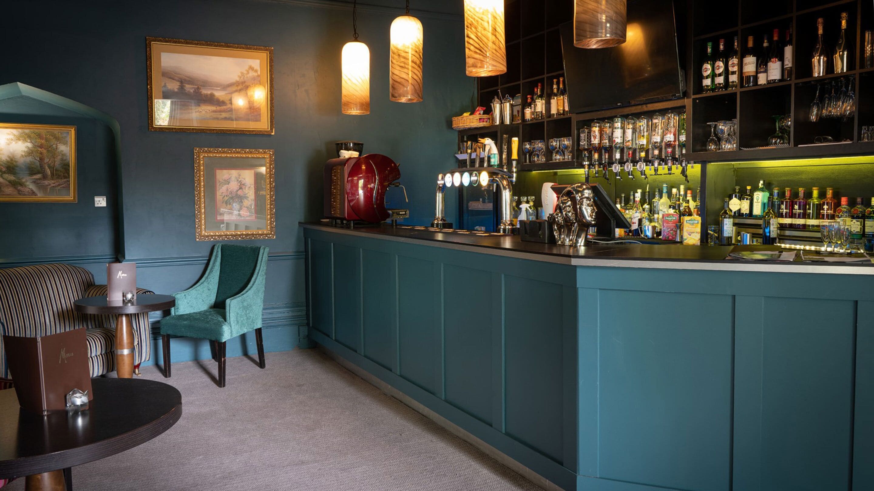 A bar area with teal cabinetry, a coffee machine, and a variety of liquor bottles. There are framed pictures on the wall and a seating area with a striped sofa and a chair.