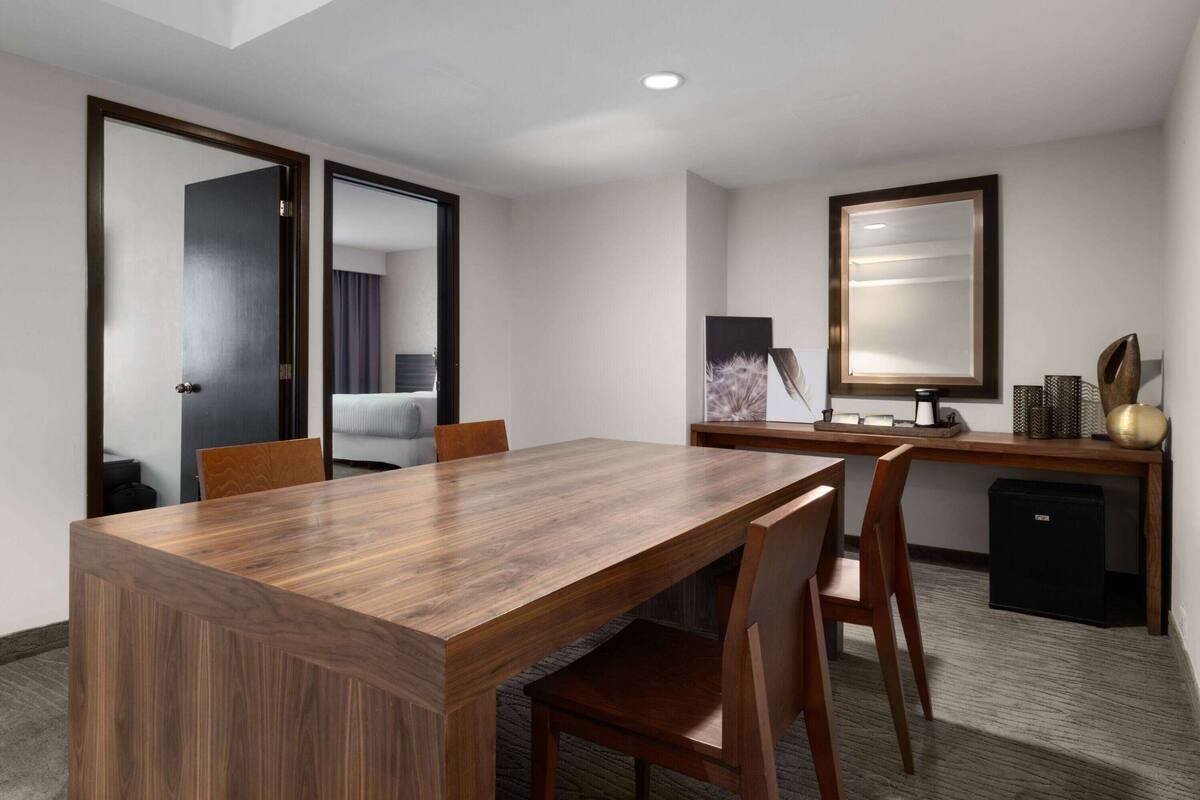 A hotel room with a wooden dining table, two chairs, a desk with a computer, a small black refrigerator, and a large mirror on the wall.