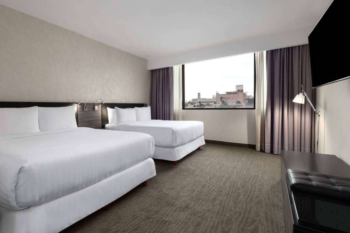 A hotel room with two beds, a desk, a large window with curtains, and a view of buildings.