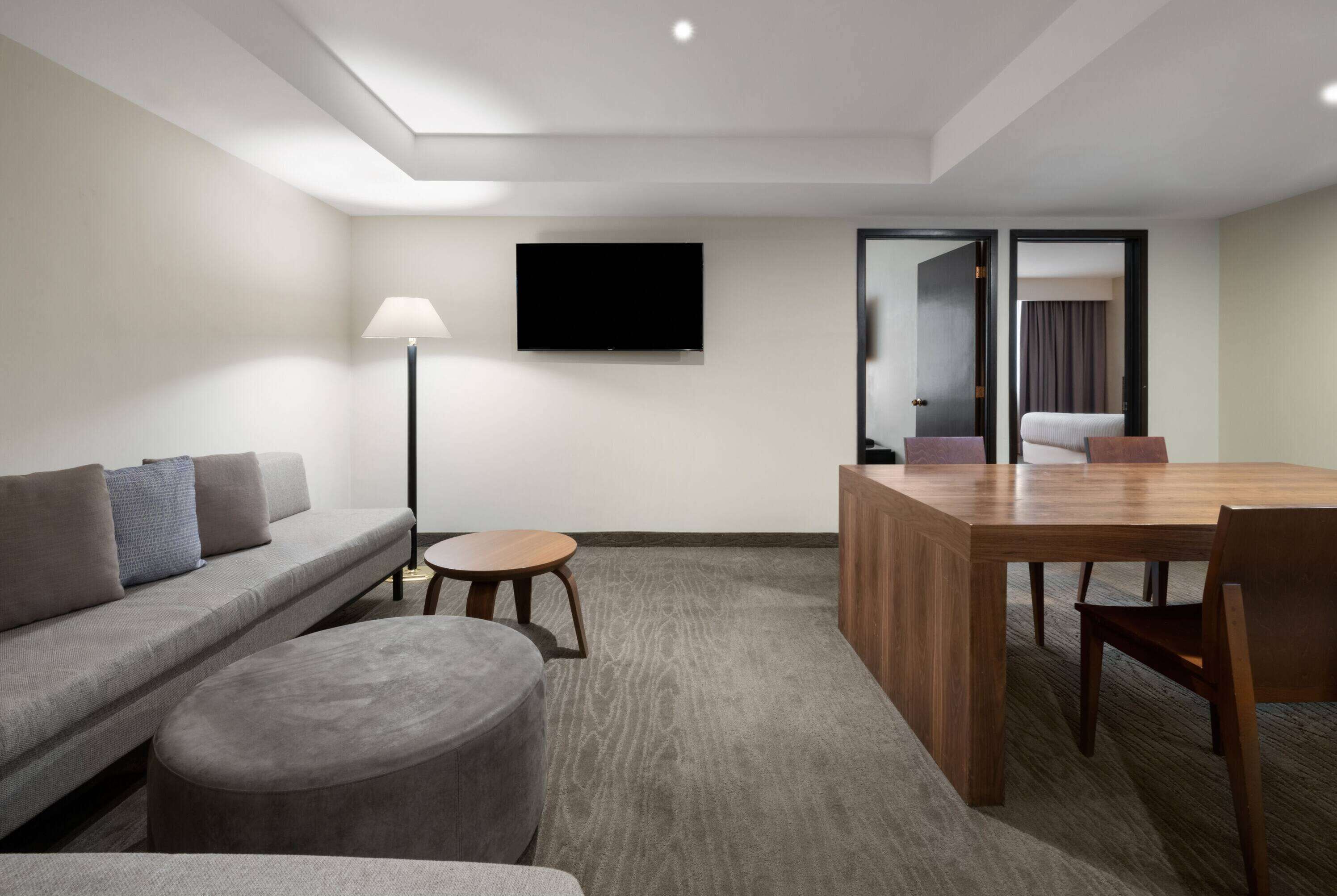 Una habitación de hotel moderna con sofá, reposapiés, una mesita redonda y una mesa de comedor de madera con sillas. Un televisor de pantalla plana está montado en la pared, y hay una puerta que conduce a otra habitación.