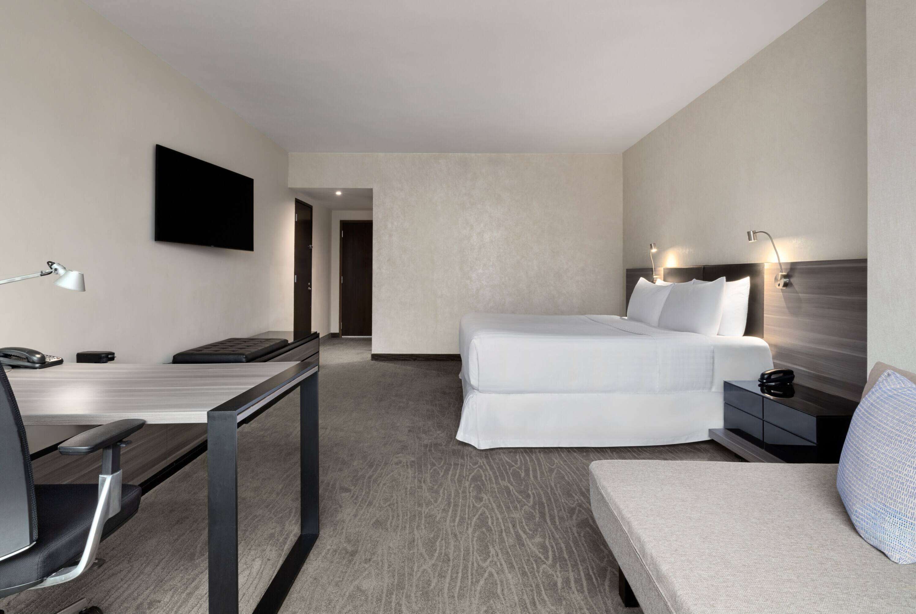 Quarto de hotel com cama, escrivaninha com cadeira, televisão na parede e mesa de cabeceira com abajur.