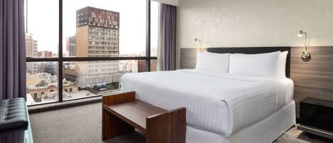 A hotel room with a large bed, a wooden bench, a view of the city through large windows, and a modern wall design.