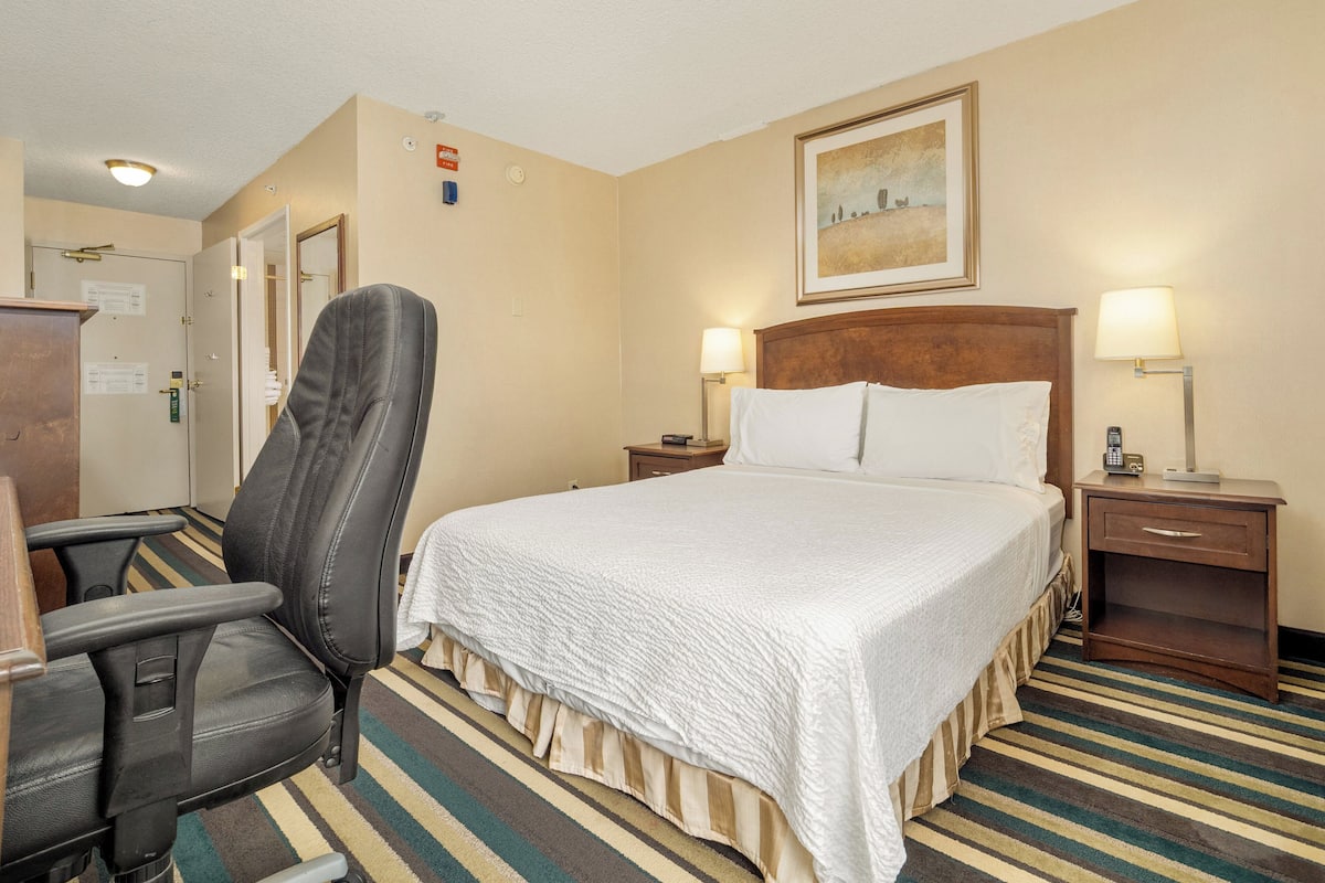 A hotel room with a bed, a chair, a desk, and a nightstand with lamps.