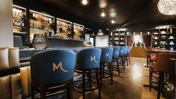 Zona de bar con taburetes altos, una barra y estanterías llenas de botellas.