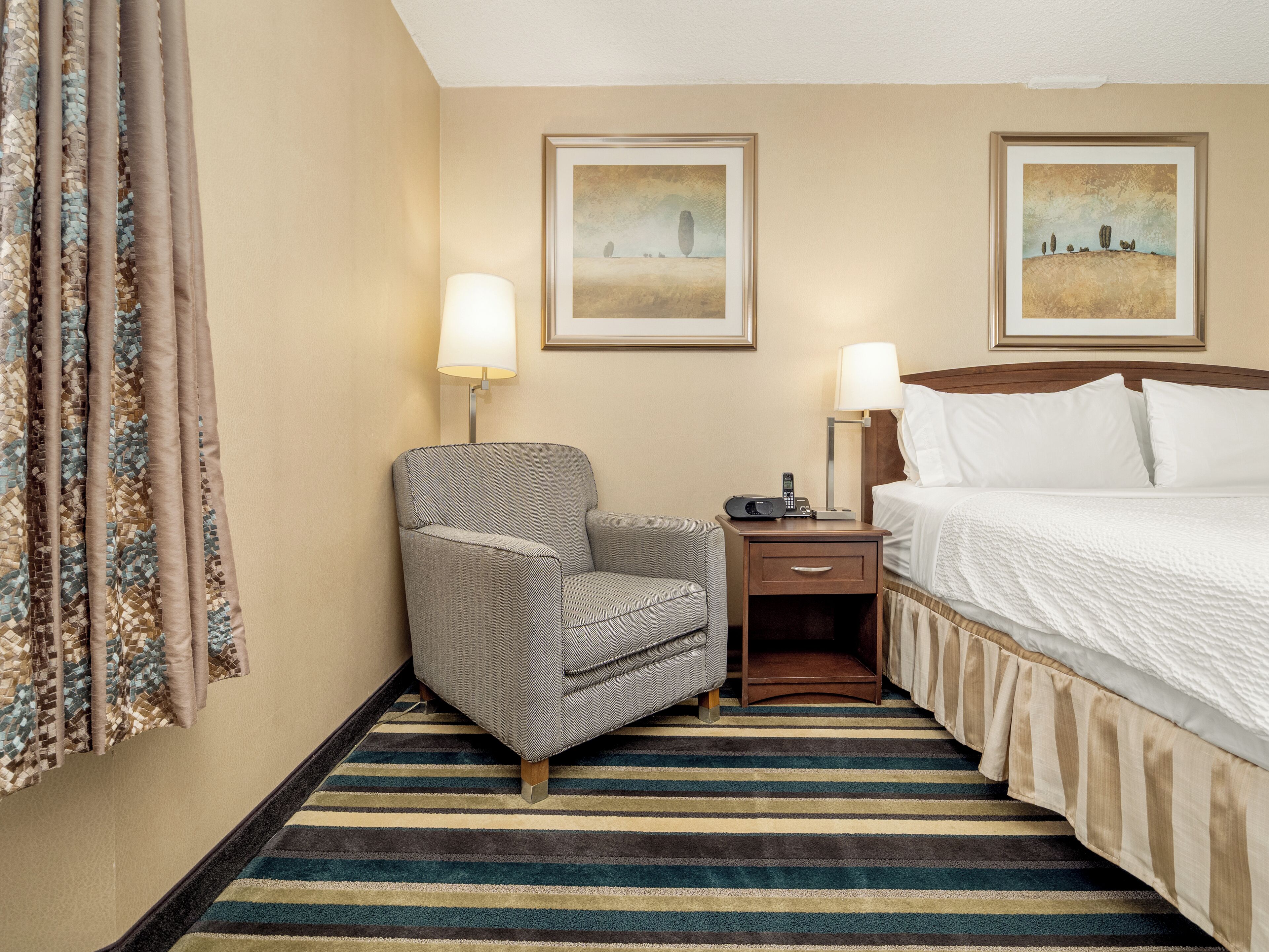Una habitación de hotel con una cama, una silla, una mesita de noche, dos lámparas y dos cuadros en la pared.