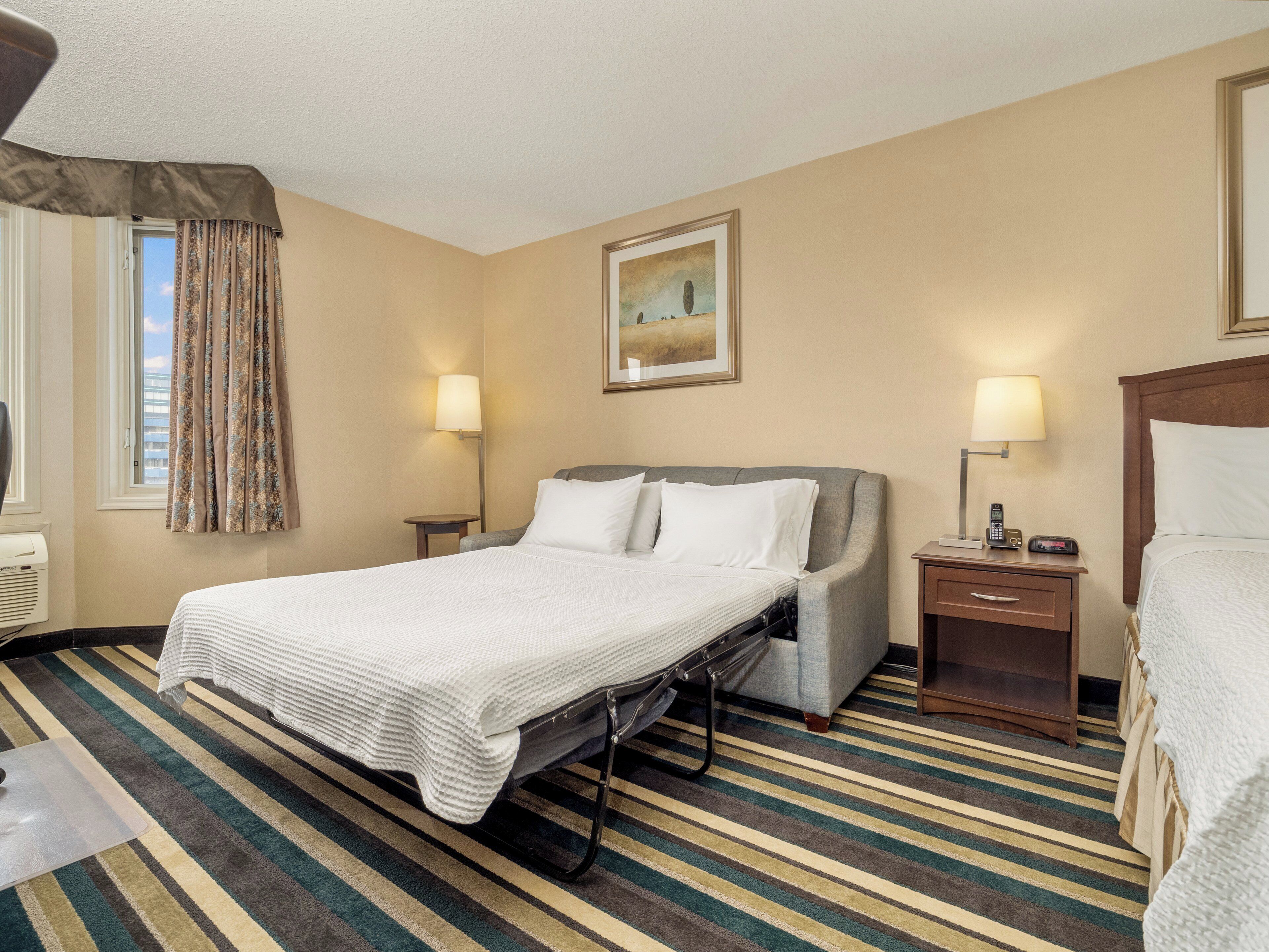 A hotel room with a bed, bedside lamps, a window with curtains, a painting on the wall, and a striped carpet.