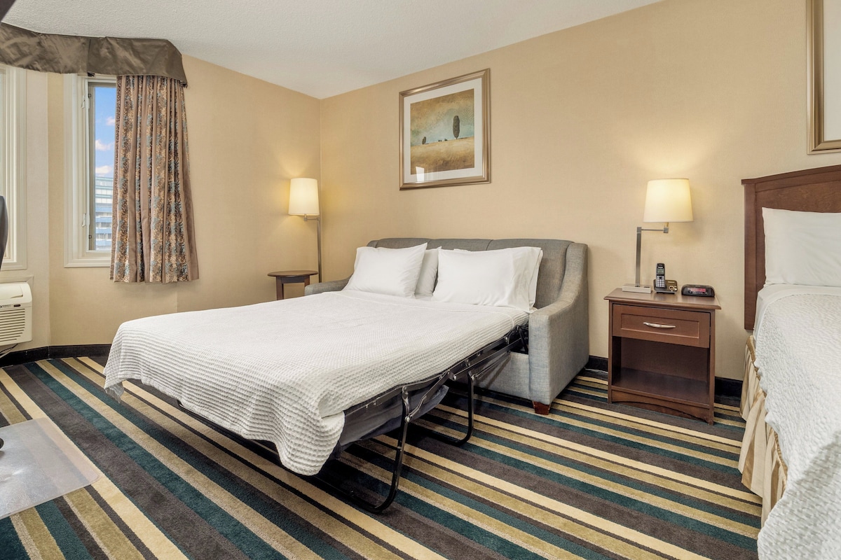 Une chambre d’hôtel avec un lit, des lampes de chevet, une fenêtre avec des rideaux, un tableau au mur et un tapis à rayures.