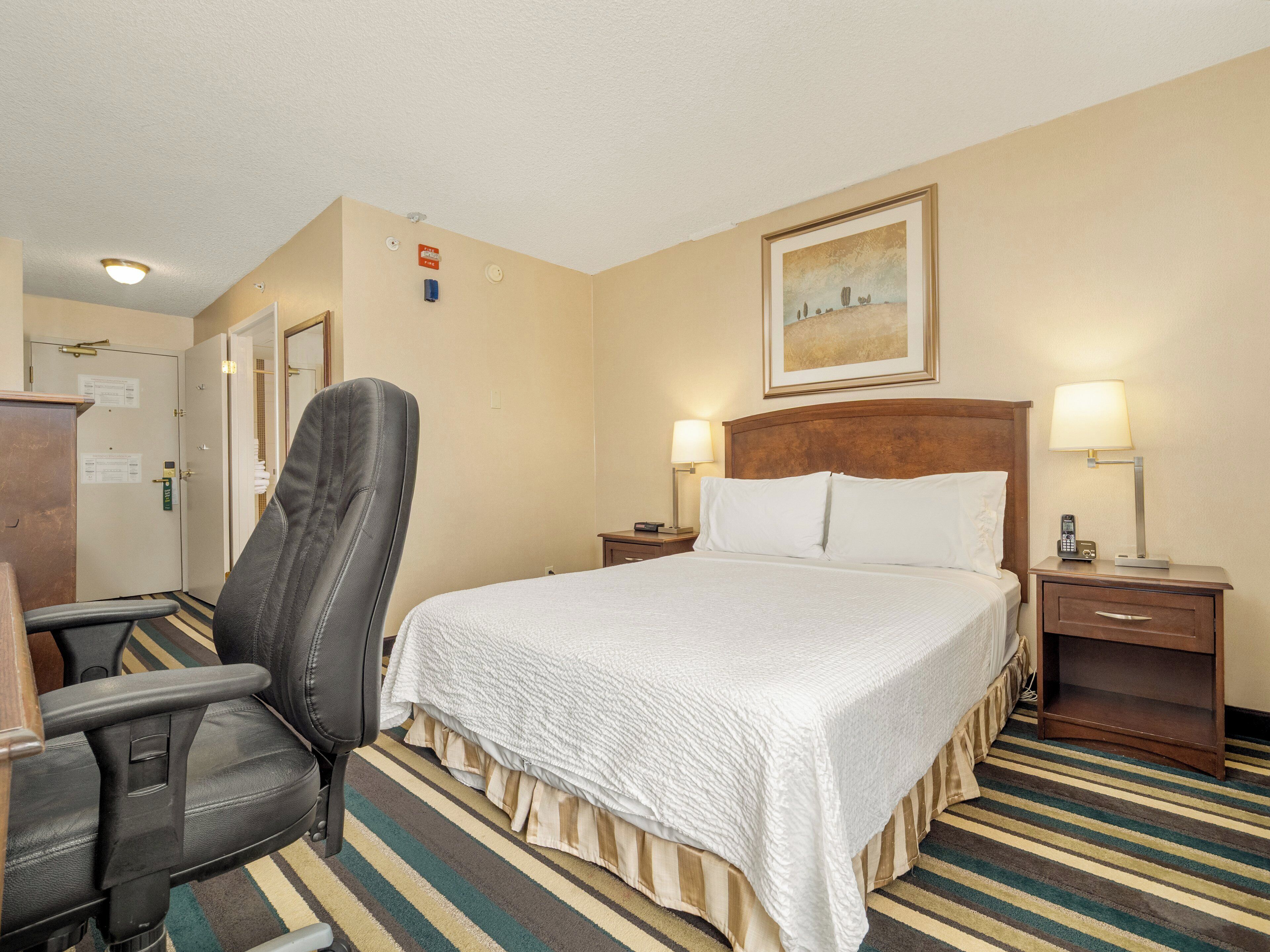 A hotel room with a bed, a chair, a desk, and a nightstand with lamps.