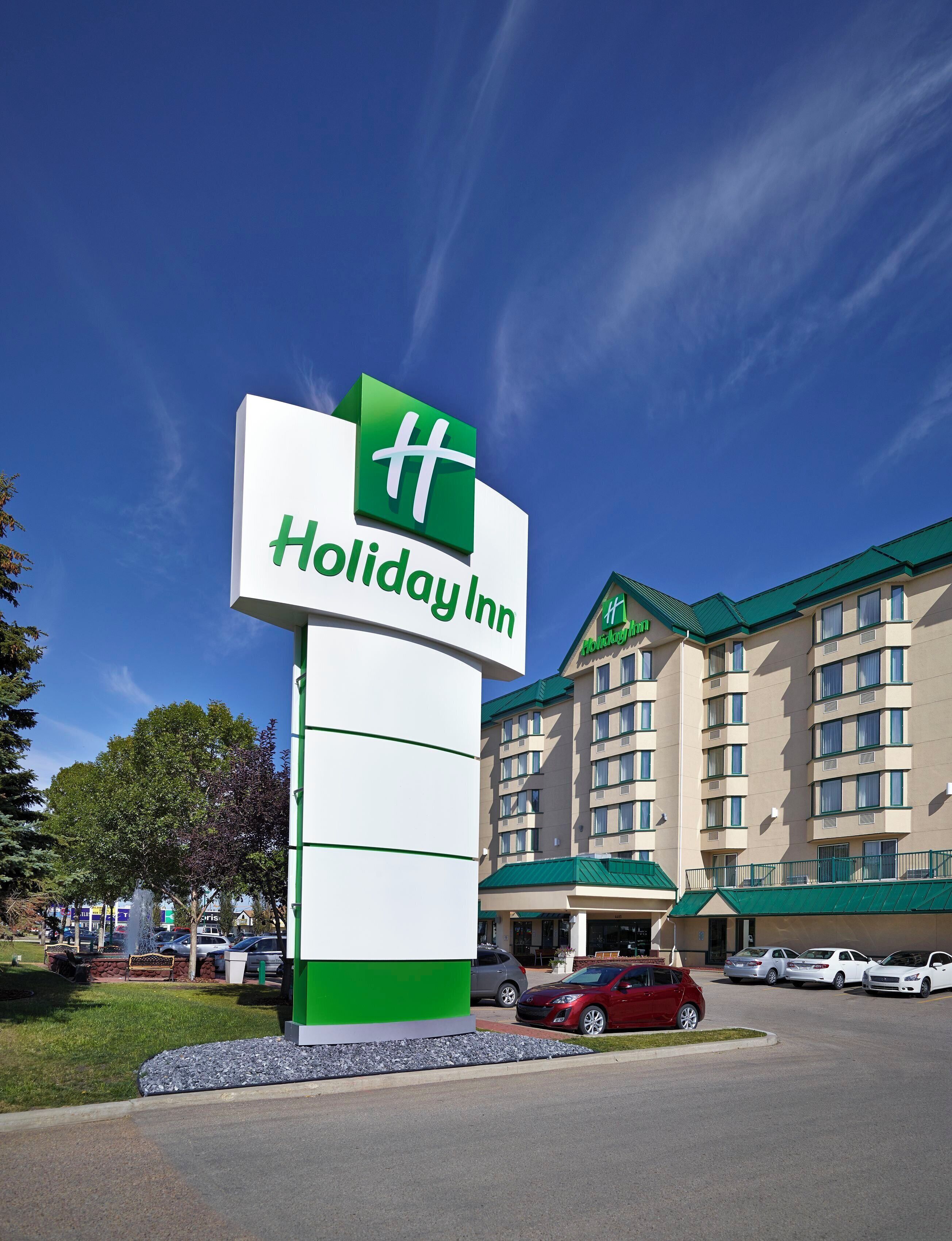 A Holiday Inn sign with a green and white logo, a parking lot with several cars, and a hotel building with multiple floors and a green roof.