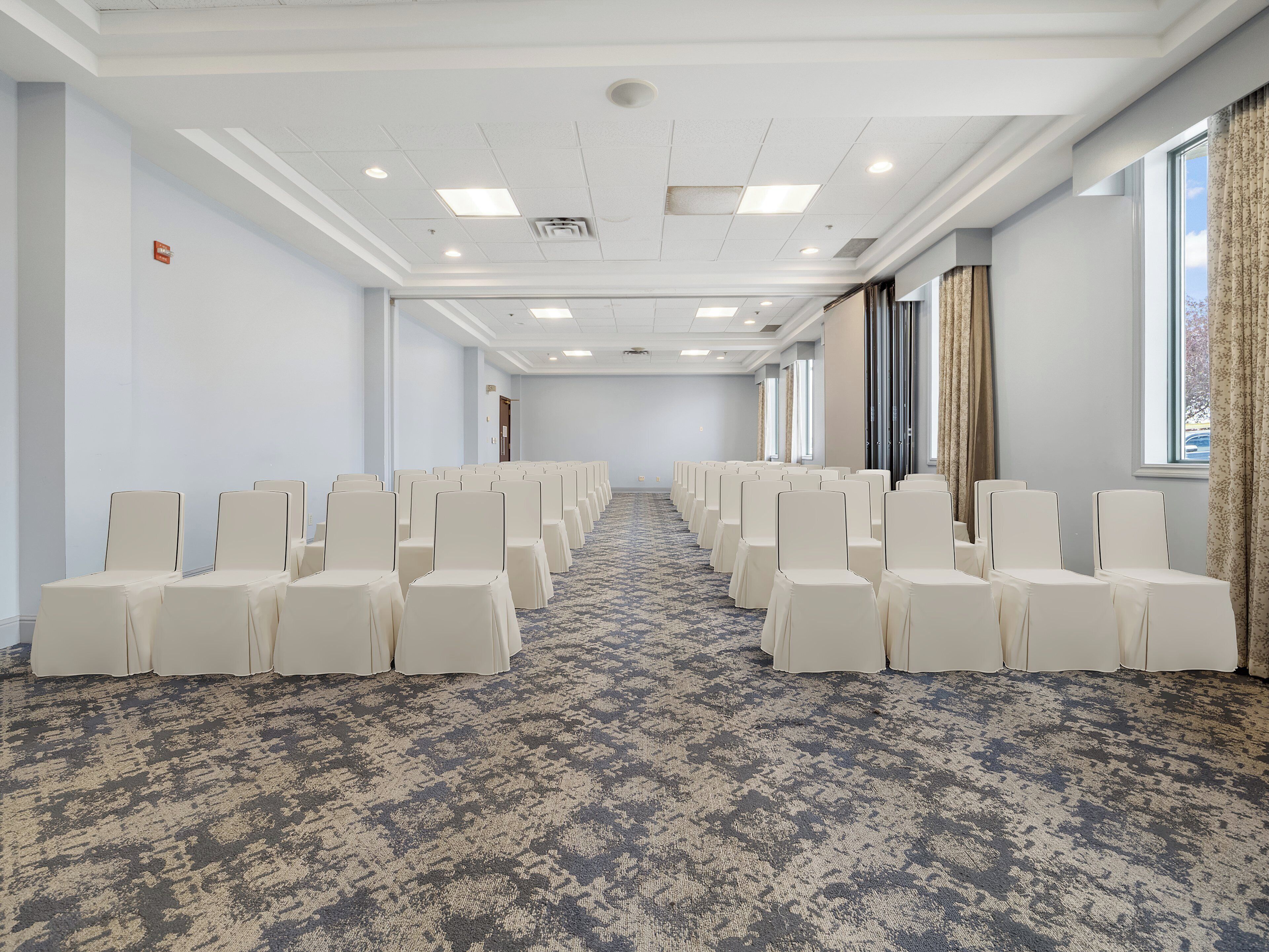 Ein Konferenzraum mit Reihen weißer Stühle, einem gemusterten Teppich und großen Fenstern mit Vorhängen.